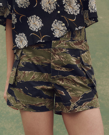 A person wears THE GREAT. SU25 The Cargo Fatigue Short in Camo Surplus and a black shirt with white floral pattern—an on-trend men’s look shown from waist to mid-thigh.