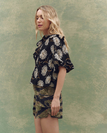 A woman with long blonde hair stands in profile, modeling THE GREAT. SU25 The Bower Top in Black Dancing Floral—a chic black floral blouse—set against a mottled green backdrop. This description enhances SEO for stylish floral tops.