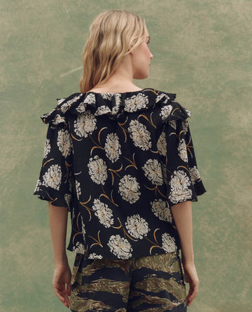 A person faces away, wearing THE GREAT. SU25's The Bower Top in Black Dancing Floral—a ruffle blouse—styled with camouflage pants against a muted green background for SEO-optimized product visibility.