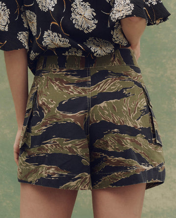Model wears The Cargo Fatigue Short—Camo Surplus by THE GREAT. SU25, featuring green and brown camo with side pockets, paired with a black floral top, photographed from the back against a green background.