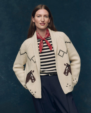 A person wears THE GREAT. SP25 COLLECTORS The Horse Lodge Cardigan in cream over a black-and-white striped shirt, paired with a red bandana and dark pants, set against a dark blue background.