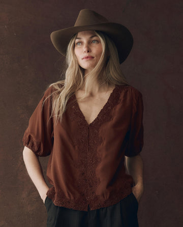 A woman with long blonde hair wears THE GREAT. F25 D1’s The Short Sleeve Legacy Top in Chocolate and a wide-brimmed brown hat, standing against a dark brown background with her hands in her pockets.