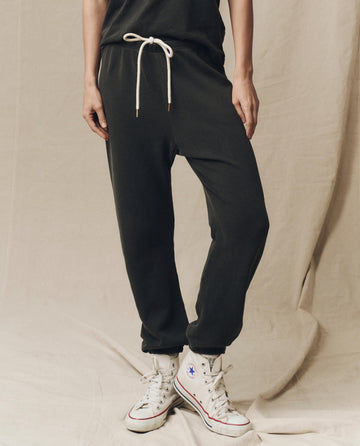 Person wearing The Stadium Sweatpant in washed black from THE GREAT. CORE KNITS with a white drawstring and high-top sneakers, effortlessly blending vintage athletic style against a beige fabric background.