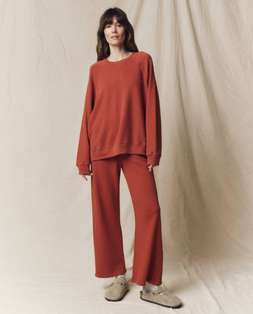 A woman stands against a beige fabric backdrop wearing THE GREAT. F25 SEASONAL KNITS The Slouch Sweatshirt, Solid -- Brick, with matching wide-leg pants and beige shoes.