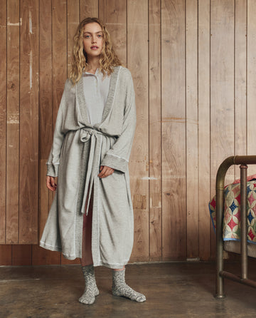 A woman stands indoors on a concrete floor, wrapped in THE GREAT. H25 SLEEP The Sweatshirt Robe in Light Heather Grey, with gray socks and a light shirt, as a patchwork quilt rests on a brass bed beside her.