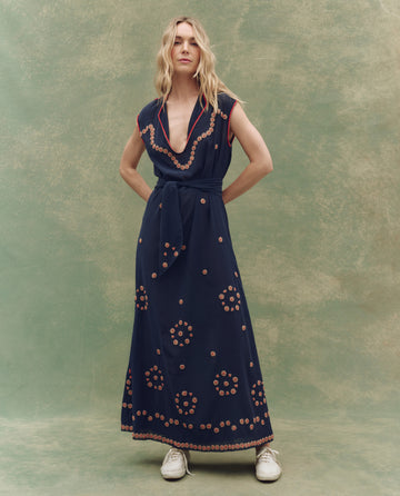 A woman stands against a mottled green backdrop wearing THE GREAT. SU25's The Beaded Corridor Dress in navy, featuring circular orange patterns, a tied waist, and white sneakers.