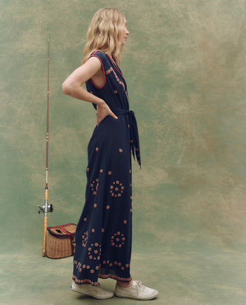 A woman in THE GREAT. SU25 The Beaded Corridor Dress – Navy, paired with white shoes, stands in profile by a fishing rod and basket against a green textured backdrop.