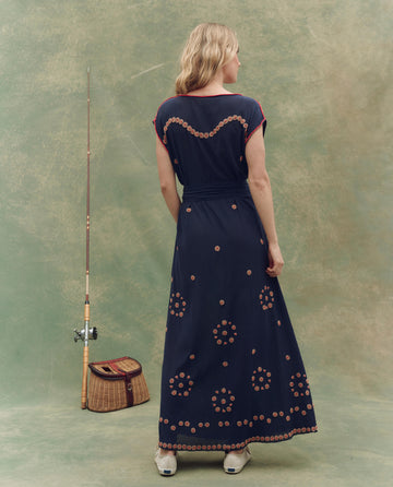A woman, back to the camera, wears THE GREAT. SU25 The Beaded Corridor Dress in navy. A fishing pole and wicker basket rest to her left against a green backdrop with circular beaded patterns visible on the dress.