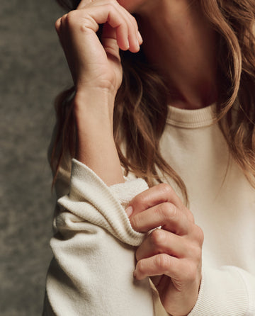 Close-up of hands rolling up the sleeve of THE GREAT. H25 CORE FLEECEBACK The Fleece College Sweatshirt in Washed White, set against a neutral background.
