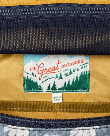 A detailed view of The Adventure Hip Pack tag reveals "THE GREAT. SP25 TGO" with a mountain and forest motif, embodying the adventure ethos. Below it, a "Made in China" label is visible.