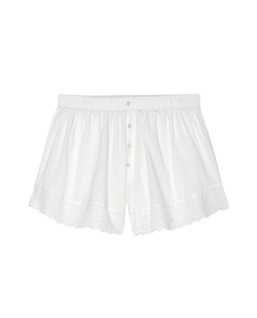 The Eyelet Tap Short in True White by THE GREAT. PS25 SLEEP, features a lightweight cotton voile material with scalloped hem, eyelet lace trim, elastic waistband, and button front closure, displayed on a black background.