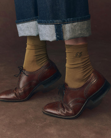 Someone, in cuffed jeans and brown leather oxfords, stands on a brown surface wearing THE GREAT. F25 D1 ACCESSORIES The Monogrammed Trouser Sock in Golden Ochre—crafted from mercerized cotton and featuring a monogram detail.