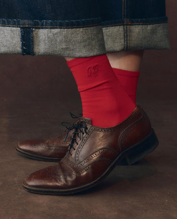 A person wears THE GREAT. F25 D1 ACCESSORIES The Monogrammed Trouser Sock in Poppy Red with monogrammed initials, paired with brown leather brogues and rolled-up denim jeans, standing on a dark floor.