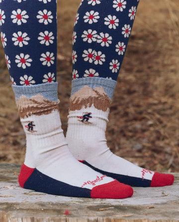Wearing THE GREAT. F25 TGO’s The Ridge Runner Sock – Cream Skiier, featuring a mountain landscape and cushioned footbed, a person stands outdoors on a tree stump in blue leggings with white and red daisy patterns.