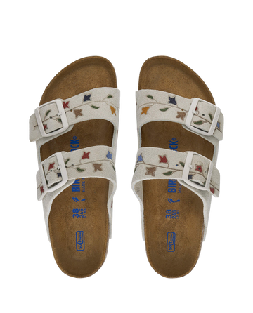 The BIRKENSTOCK Arizona by THE GREAT. SU24 BIRKENSTOCK features hand-painted, tooled tulip patterns. With elegant antique white straps adorned with multicolored leaves, the sandals offer two buckles and a comfy brown cork footbed.