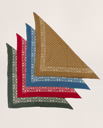 Five ultra-soft triangular bandanas from THE GREAT. SP24 D2's collection, in olive green, red, blue, brown, and dark green with a distinctive white floral pattern along the edges, are neatly laid out in an overlapping arrangement on a white background.