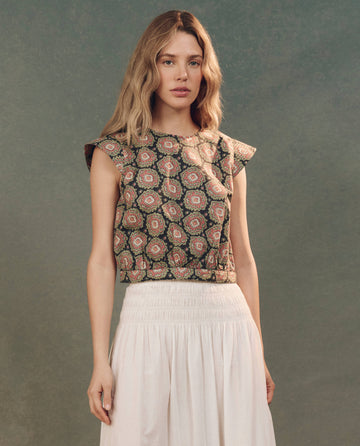 A person with long hair wears The Midland Top in Heirloom Paisley by THE GREAT. SP25 D2, paired with a white skirt, standing against a textured background.