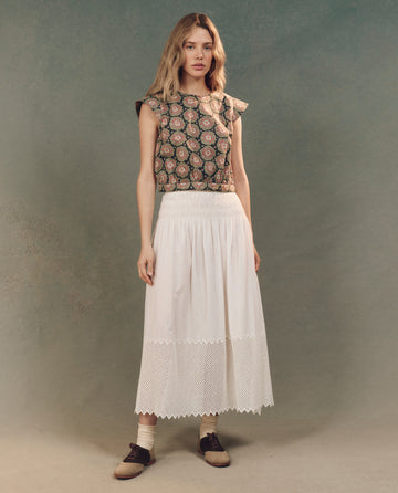 Against a gray background, the woman showcases elegant attire with fashion-forward flair, wearing The Midland Top in Heirloom Paisley by THE GREAT. SP25 D2, paired with a long white skirt with lace trim, complemented by brown shoes and white socks.