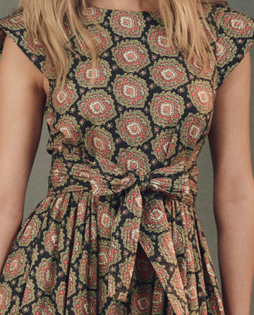 The Tulsa Dress by THE GREAT. SP25 D2 features a vibrant sleeveless design with an intricate red and green Heirloom Paisley pattern, highlighted by a large waist bow, epitomizing elegance in women's clothing.