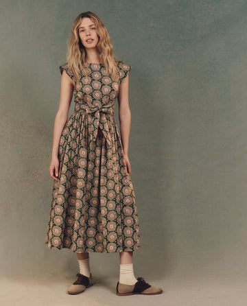 A woman poses against a textured backdrop, dressed in The Tulsa Dress - Heirloom Paisley by THE GREAT. SP25 D2, paired with vintage-style shoes and socks.