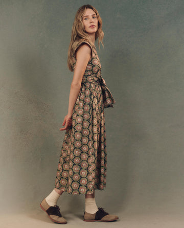 A woman stands sideways in THE GREAT.'s The Tulsa Dress -- Heirloom Paisley, elegantly paired with white socks and dark shoes against a muted backdrop.