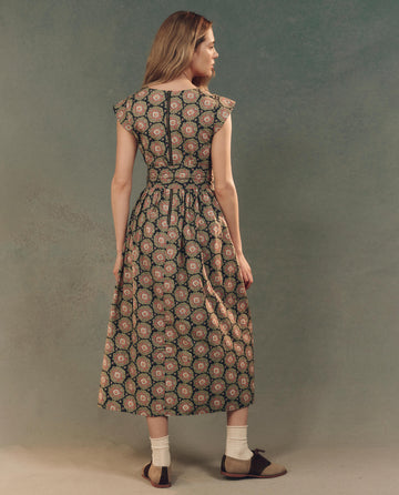A woman stands facing away in "The Tulsa Dress" by THE GREAT. SP25 D2, featuring an Heirloom Paisley pattern. She pairs it with ankle socks and black flats against a textured green backdrop.