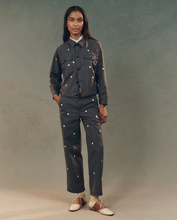 A person stands against a muted gray background, wearing THE GREAT. SP25 D2's The Garage Jacket with Daisy Embroidery in Blue Slate, paired with trousers and brown and white shoes.