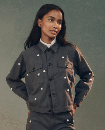 A person in THE GREAT. SP25 D2's The Garage Jacket with Daisy Embroidery in Blue Slate and matching pants stands against a muted background.