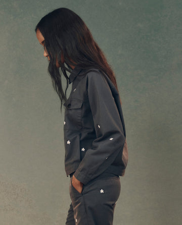 A person in profile wears THE GREAT.'s Garage Jacket with Daisy Embroidery in Blue Slate, set against a textured green background.