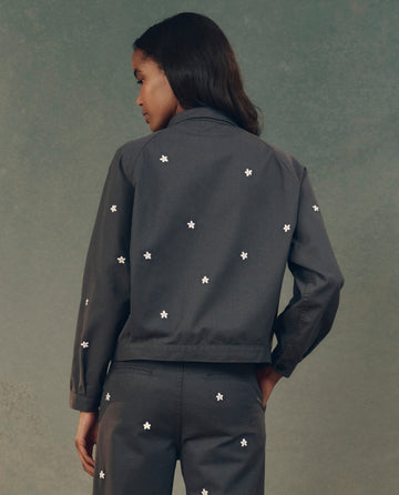 A person stands facing away against a muted background, wearing THE GREAT. SP25 D2's The Garage Jacket with Daisy Embroidery in Blue Slate paired with matching pants showcasing delicate white daisy embroidery.