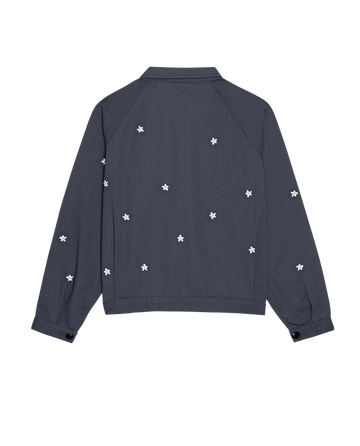 The Garage Jacket with Daisy Embroidery by THE GREAT. SP25 D2 in Blue Slate showcases a dark blue tone with delicate white flower patterns on the back, daisy embroidery on long sleeves, buttoned cuffs, and a classic collar.