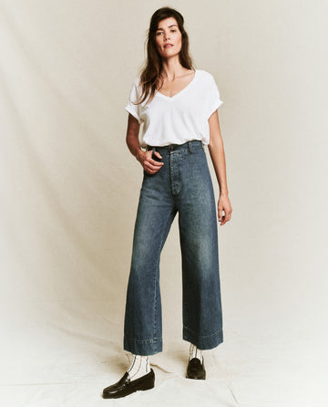 A person wearing a white t-shirt, The Seafair Jean by THE GREAT. PF24 in Marina Wash with wide legs, and black shoes stands against a plain background.