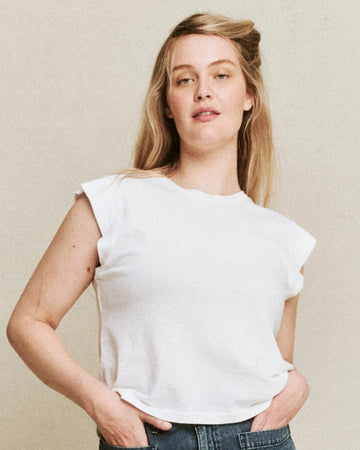 A woman with long blonde hair wearing a handcrafted locally The Peak Shoulder Tee by THE GREAT. FALL 22 KNITS in True White and jeans is standing with her hands in her pockets.