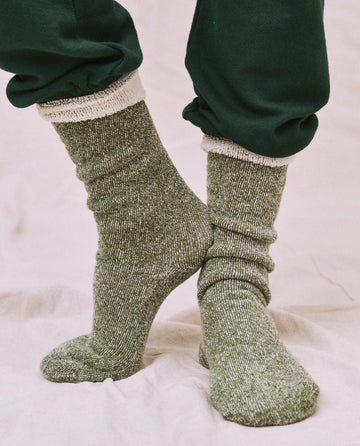 A person wearing THE GREAT. SP25 SWEATSHIRT SOCKS in Deep Pine, paired with dark green pants, stands on a beige surface, highlighting their casual footwear style.