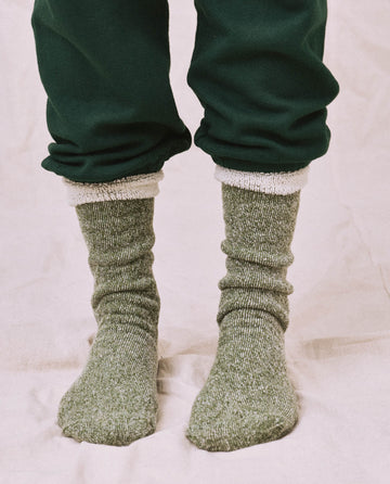 An individual wearing THE GREAT. SP25 Sweatshirt Sock in Deep Pine and dark green pants stands confidently on a light-colored surface.