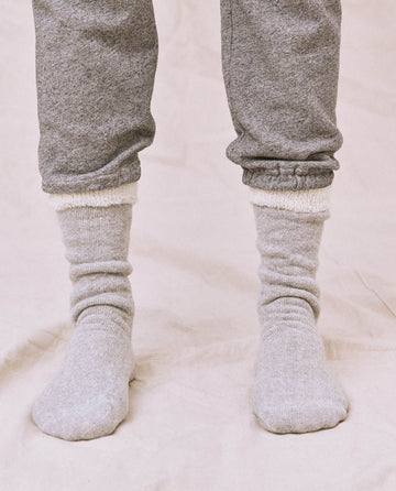 A person in THE GREAT. SP25 Sweatshirt Socks - Heather Grey stands on a soft fabric surface, wearing cozy gray sweatpants and a matching sweatshirt.
