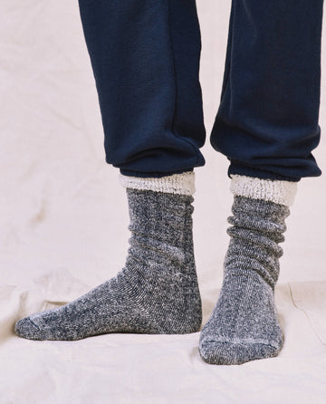 A stylish individual wears THE GREAT. SP25 Sweatshirt Sock—a gray marled design with white cuffs—paired perfectly with navy blue pants for an effortlessly chic fashion statement.
