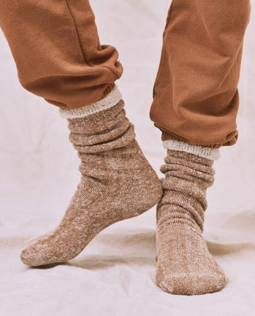 A person in brown sweatpants and a matching cozy sweatshirt stands on a light-colored surface, their feet snug in THE GREAT. SP25 Sweatshirt Socks titled "The Sweatshirt Sock" in Nutmeg.