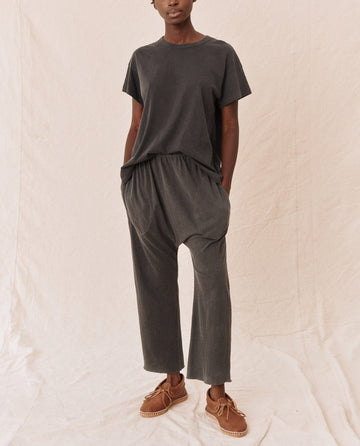 A person wearing The Jersey Crop in washed black from THE GREAT. CORE KNITS stands against a beige backdrop, hands in pockets and wearing brown shoes.
