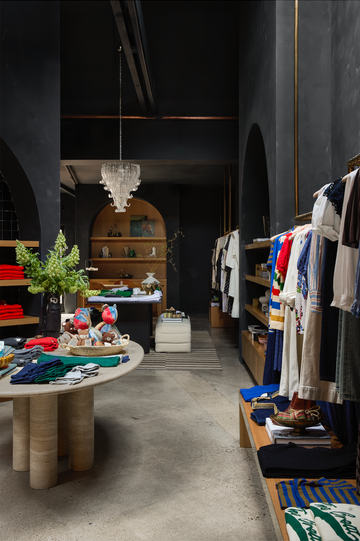 A boutique clothing store interior with neatly arranged clothes on shelves and tables, a chandelier overhead, and decorative plants placed throughout the space.
