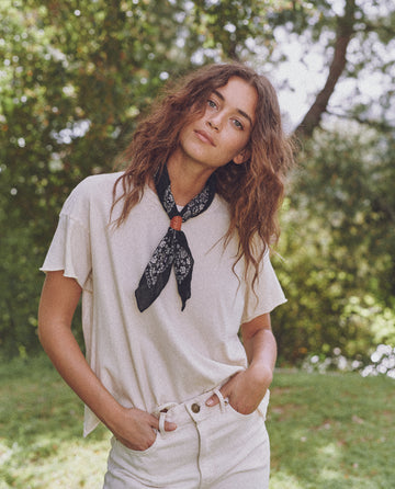 A person with curly hair wearing a white t-shirt, white pants, and "The Bandana - Small" in Almost Black by THE GREAT. SP24 D2 stands outdoors with greenery in the background.