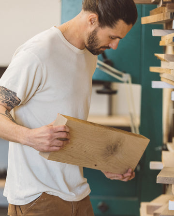 A man with a tattoo on his left arm examines a wooden plank in a workshop. He is wearing The Men's Boxy Crew in Washed White by THE GREAT. MAN, along with brown pants.
