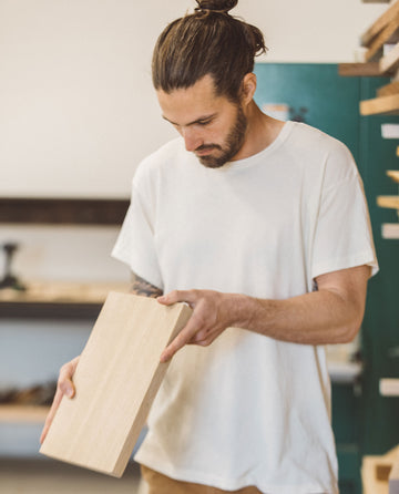 A person in The Men's Boxy Crew in Washed White by THE GREAT. MAN inspects a wooden plank in a workshop environment.