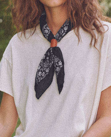 A person with long hair wearing a white t-shirt and The Bandana. -- Small -- Almost Black from THE GREAT. SP24 D2 around their neck, reminiscent of vintage handkerchiefs.