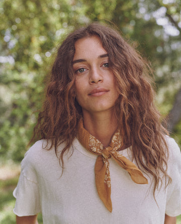 A woman with wavy hair, wearing a white shirt and a brown scarf, stands outdoors with greenery in the background. Her scarf, The Bandana - Small - Marigold by THE GREAT. SP24 D2, made from ultra-soft 100% cotton, adds a touch of comfort and style to her outfit.