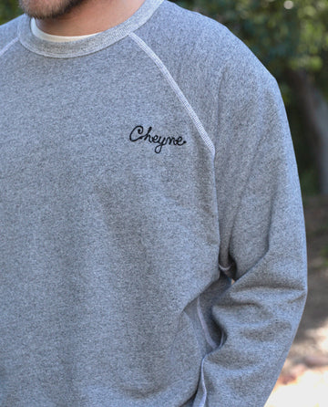 A person wearing The GREAT. MAN's Men's College Sweatshirt in Varsity Grey, with the name "Cheyne" embroidered on the left chest, stands outdoors.
