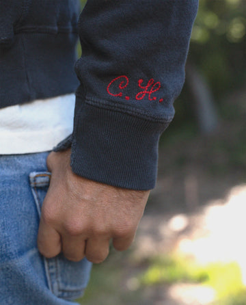 Close-up of a person's hand wearing The Men's College Sweatshirt in Washed Navy by THE GREAT. MAN., featuring dark blue vintage styling with red initials "C.H." embroidered on the sleeve, paired with blue jeans.