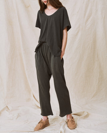 A person wearing a dark, loose-fitting casual outfit featuring The Jersey Crop in Washed Black from THE GREAT. CORE KNITS, paired with beige shoes, stands against a light fabric backdrop with hands in pockets.