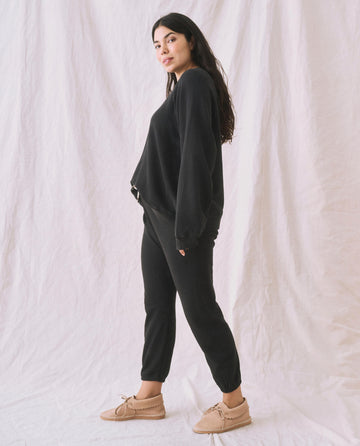 A person stands sideways in The Slouch Sweatshirt by THE GREAT. CORE KNITS, in Solid -- Almost Black, with that perfect extra slouch, paired with black pants and beige shoes, in front of a white fabric backdrop.