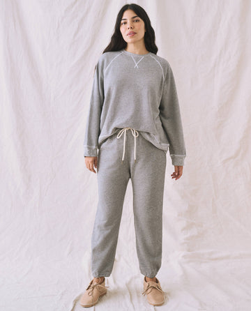 A person stands against a plain background wearing The Slouch Sweatshirt in Varsity Grey by THE GREAT. CORE KNITS, featuring fuller sleeves, along with matching sweatpants with drawstrings, and beige slip-on shoes.